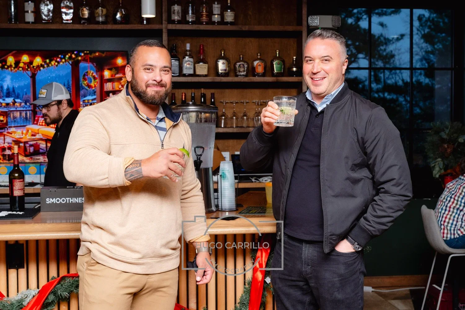 Two men smiling and holding drinks in a bar, with a bartender and holiday decorations in the background.