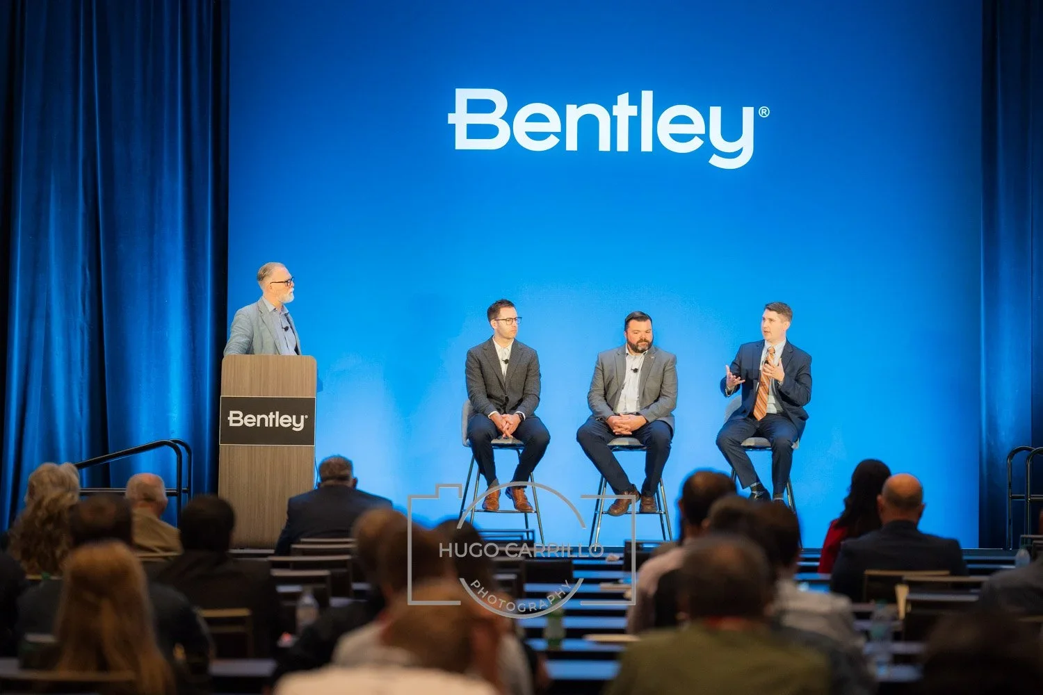 A panel discussion at a Bentley event with four men on stage, three seated and one standing at a podium, with an audience watching.