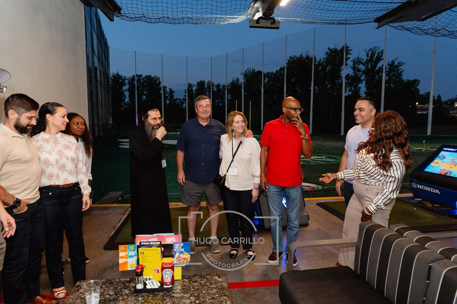 Group of nine people standing and talking inside a sports bar or golf simulation lounge during lighting evening, with golf driving range nets visible in the background.