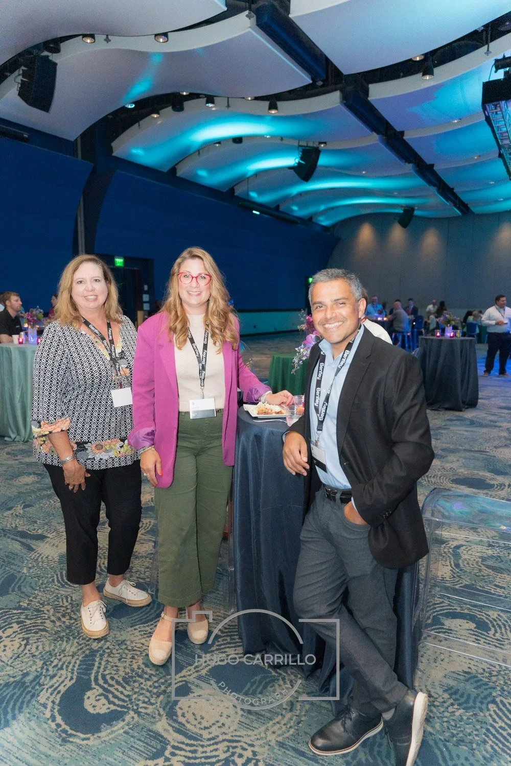 Three professionally dressed individuals at a conference or event, standing and smiling in a large, modern interior space with round tables, floral arrangements, and blue lighting.