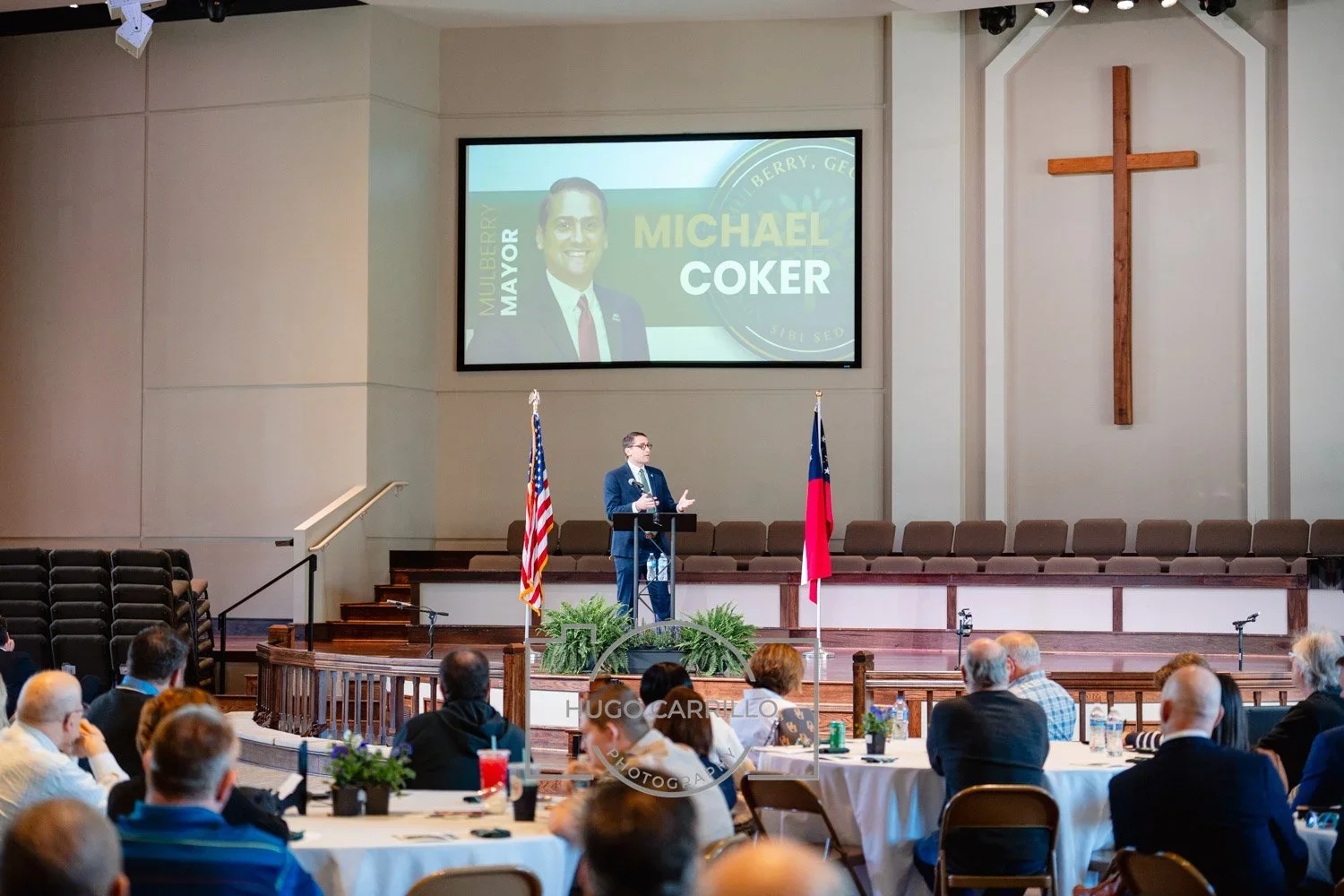 A man speaking at a podium with American and Georgia flags in front of him during a formal event in a church. Behind him is a large screen displaying a graphic with a photo and the name Michael Coker. The church interior features a wooden cross on th