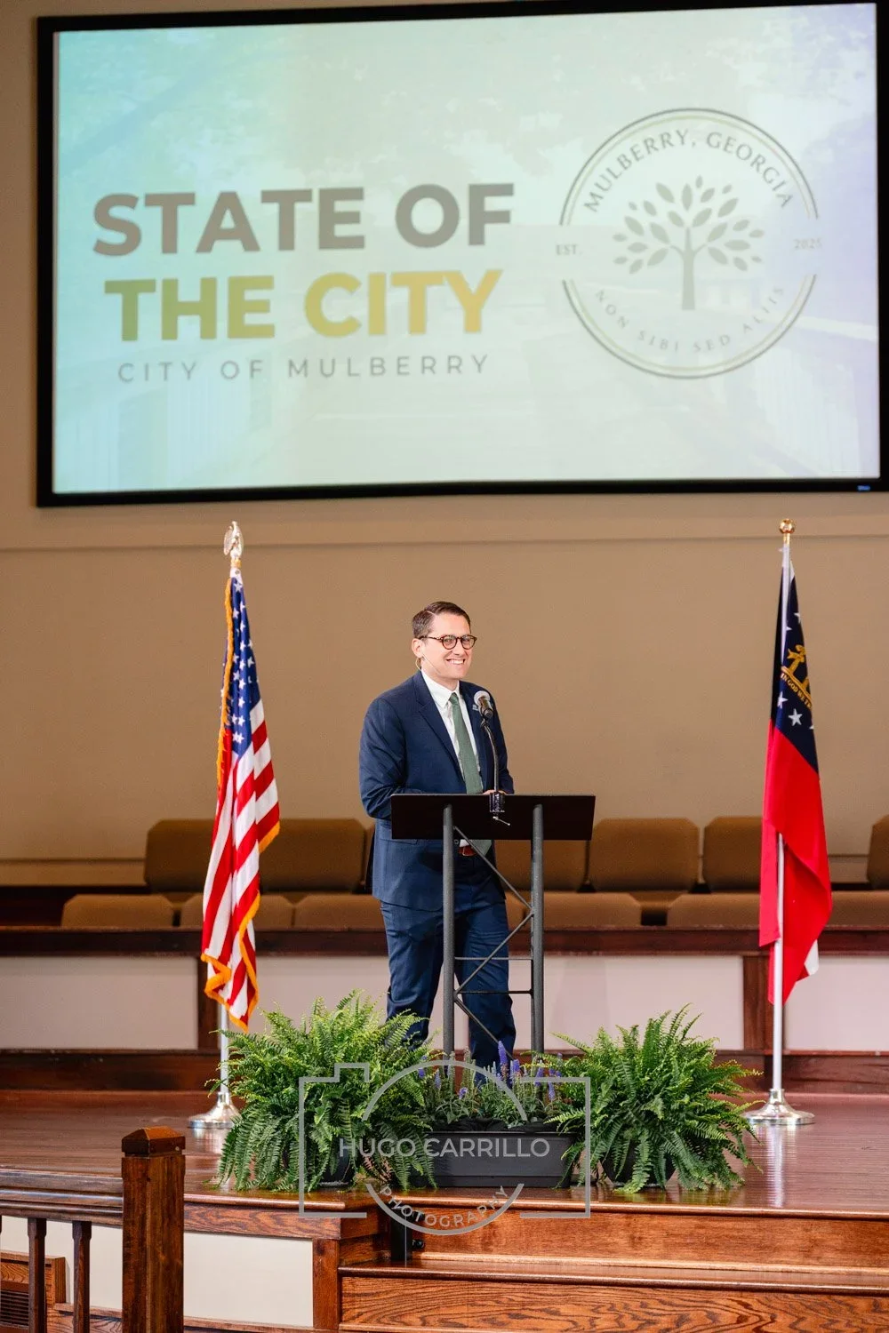 A man in a suit and glasses is standing at a podium with a microphone, smiling. Behind him are two flags, one American and one Georgia state flag, and a large screen displaying 'State of the City, City of Mulberry.'