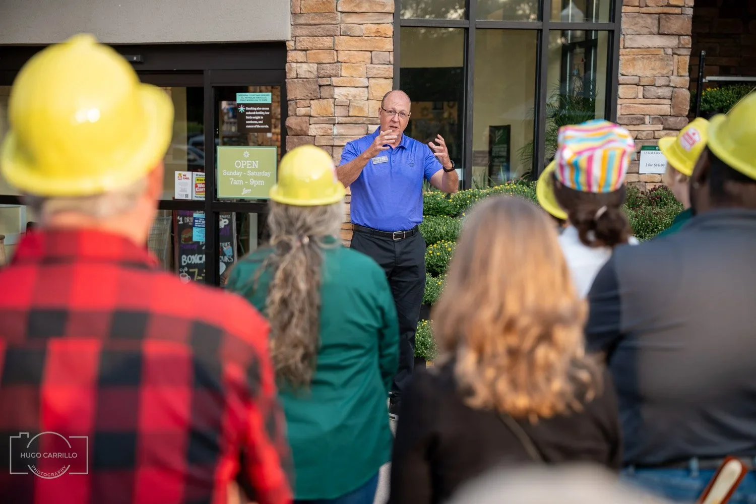 A man in a blue shirt speaking to a group wearing yellow hard hats outside a building with a sign that says "Open Sunday - Saturday 7am - 9pm." The group includes people with colorful hats and casual clothing, listening attentively.