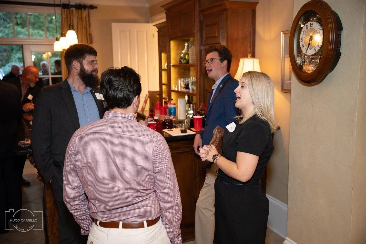 Group of four people conversing in a kitchen or dining area at a social gathering, with a bar in the background and a large wall clock on the wall.