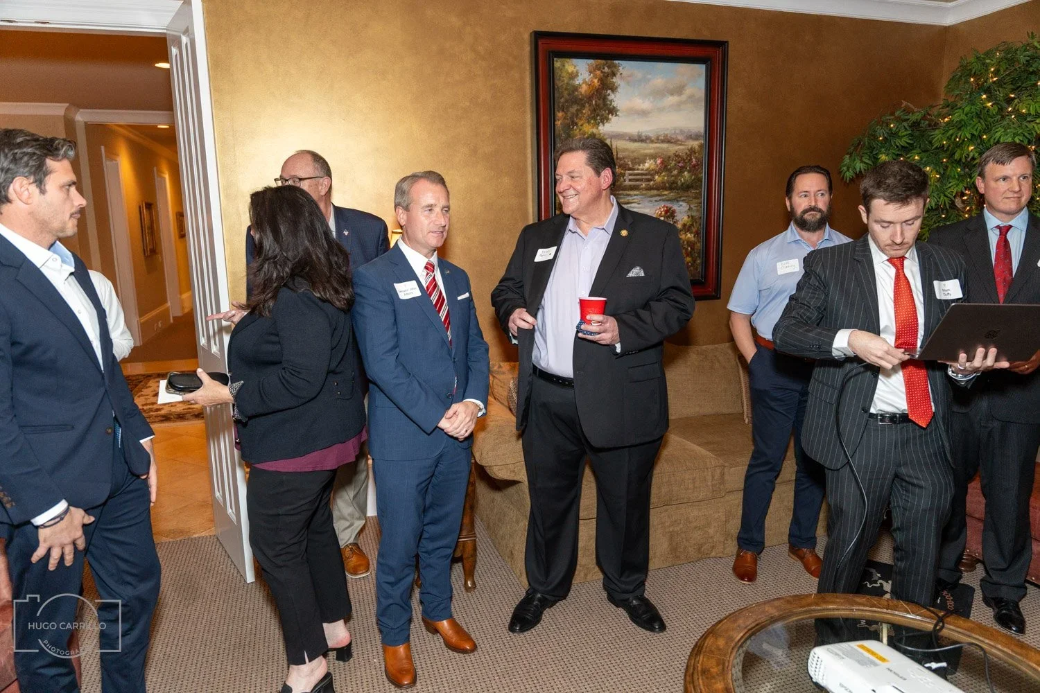 Group of people in business attire socializing at an indoor event, with a painting on the wall and a large potted plant in the background.