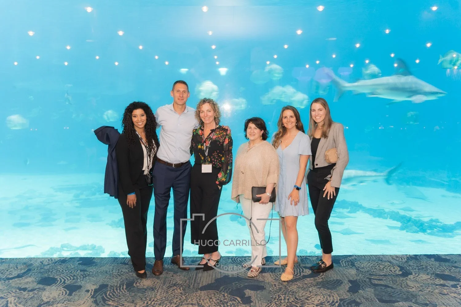 Group of six people standing in front of a large aquarium tank with fish and a shark swimming in the background.