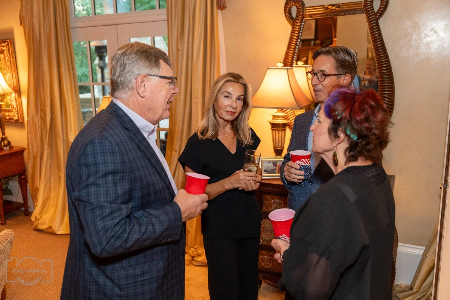 Four adults having a conversation at a social gathering, with three holding red cups and one holding a glass of water, inside a warmly lit living room.