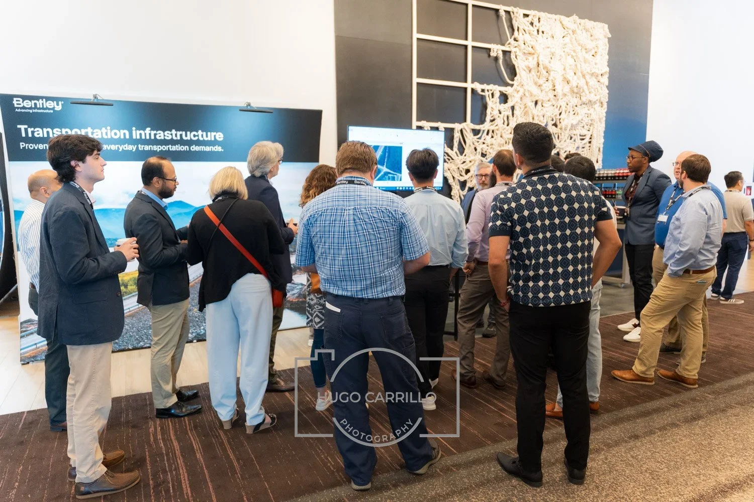 Group of people at a conference booth, listening to a presentation on transportation infrastructure, with screens and branding in the background.