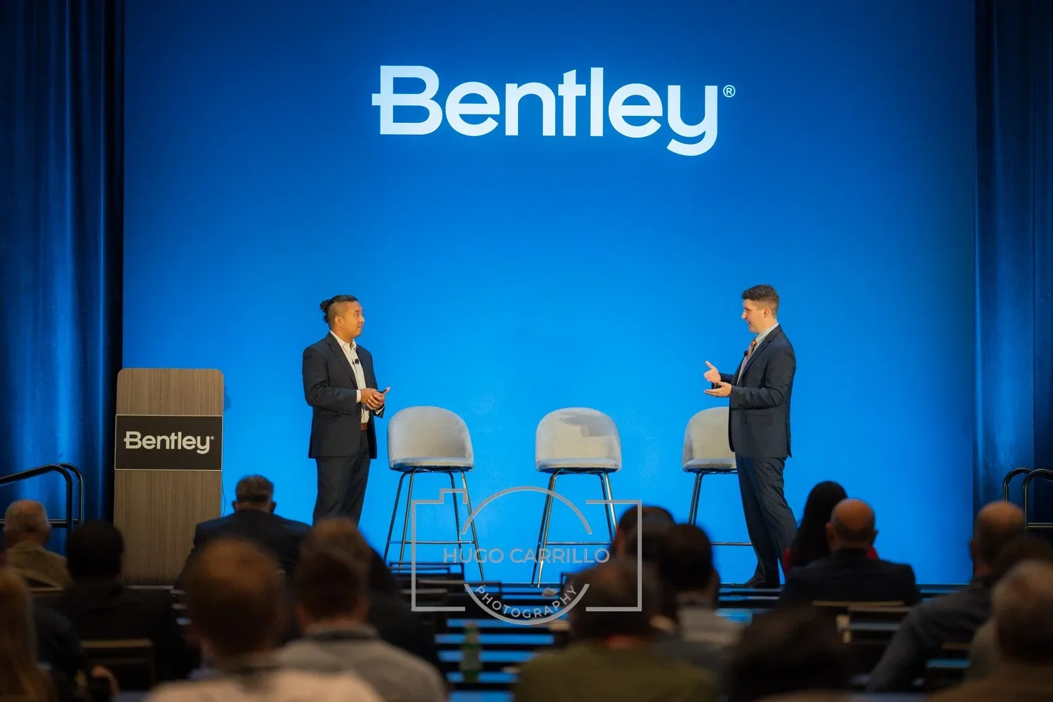 Two men in suits stand on a stage with a blue backdrop displaying the 'Bentley' logo. There are three empty chairs between them and an audience watching.