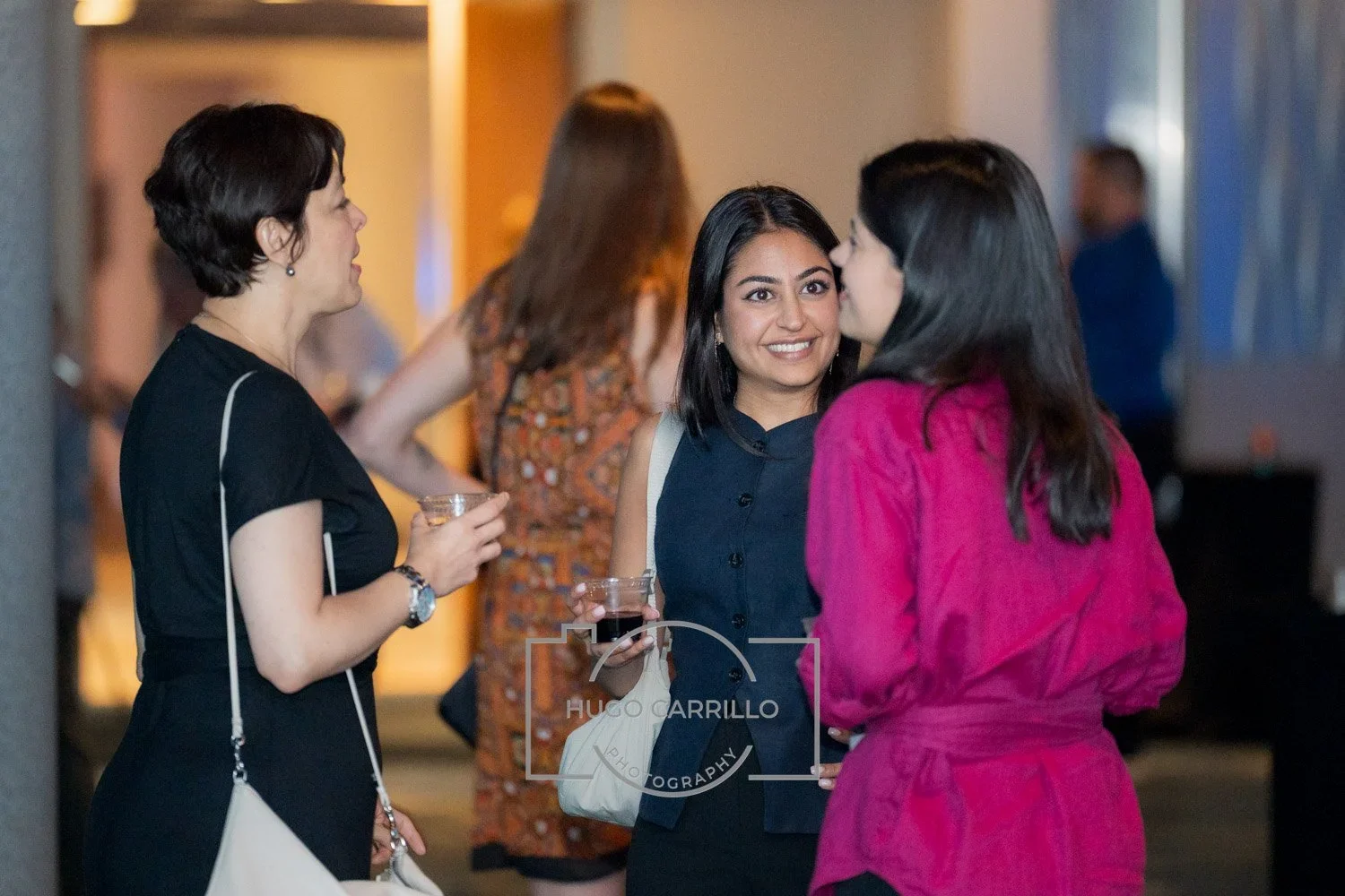Three women engaged in conversation at a social event, holding drinks, with one woman smiling at the camera.