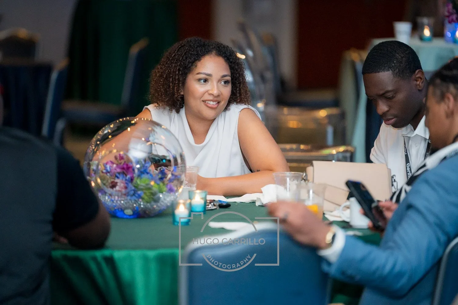 A woman with curly brown hair smiling while sitting at a round table with a group of people at a formal event or conference, with a decorative glass bowl of colorful flowers and small candles on the table.