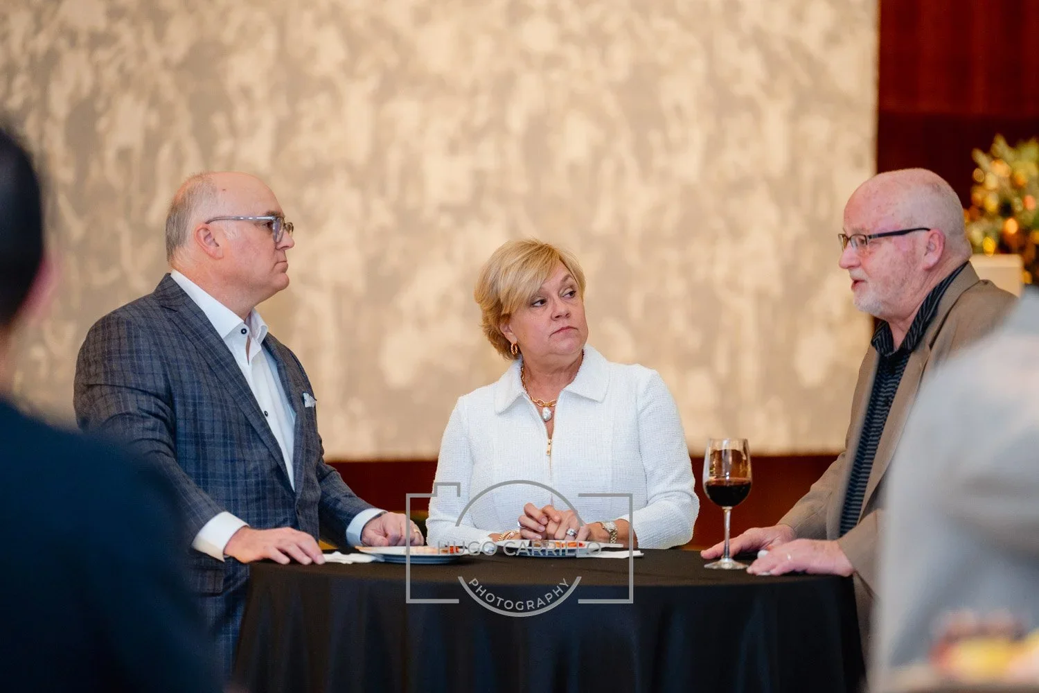 Three adults seated at a black table during a social event, with a woman in the center wearing a white jacket and gold jewelry, a man on the left in a checkered blazer, and a man on the right with a wine glass and beige blazer, engaged in conversatio