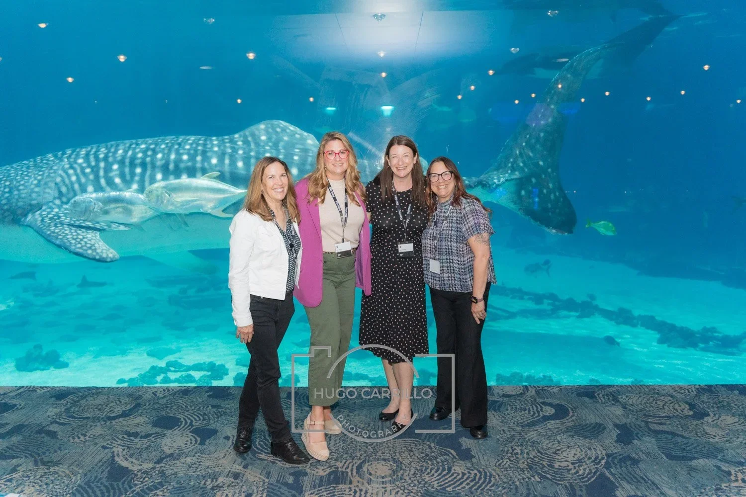 Four women standing in front of an aquarium tank with a whale shark, several fish, and marine life behind glass.