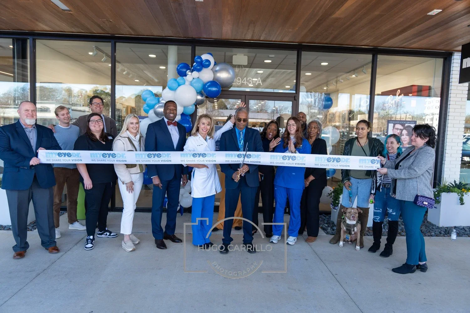 A diverse group of people gathered outside a building with glass doors for a ribbon-cutting ceremony. They are holding a ribbon that reads 'my eye dr. WE SHARE YOUR VISION.' There are balloons in blue, white, and silver behind them, and the event see