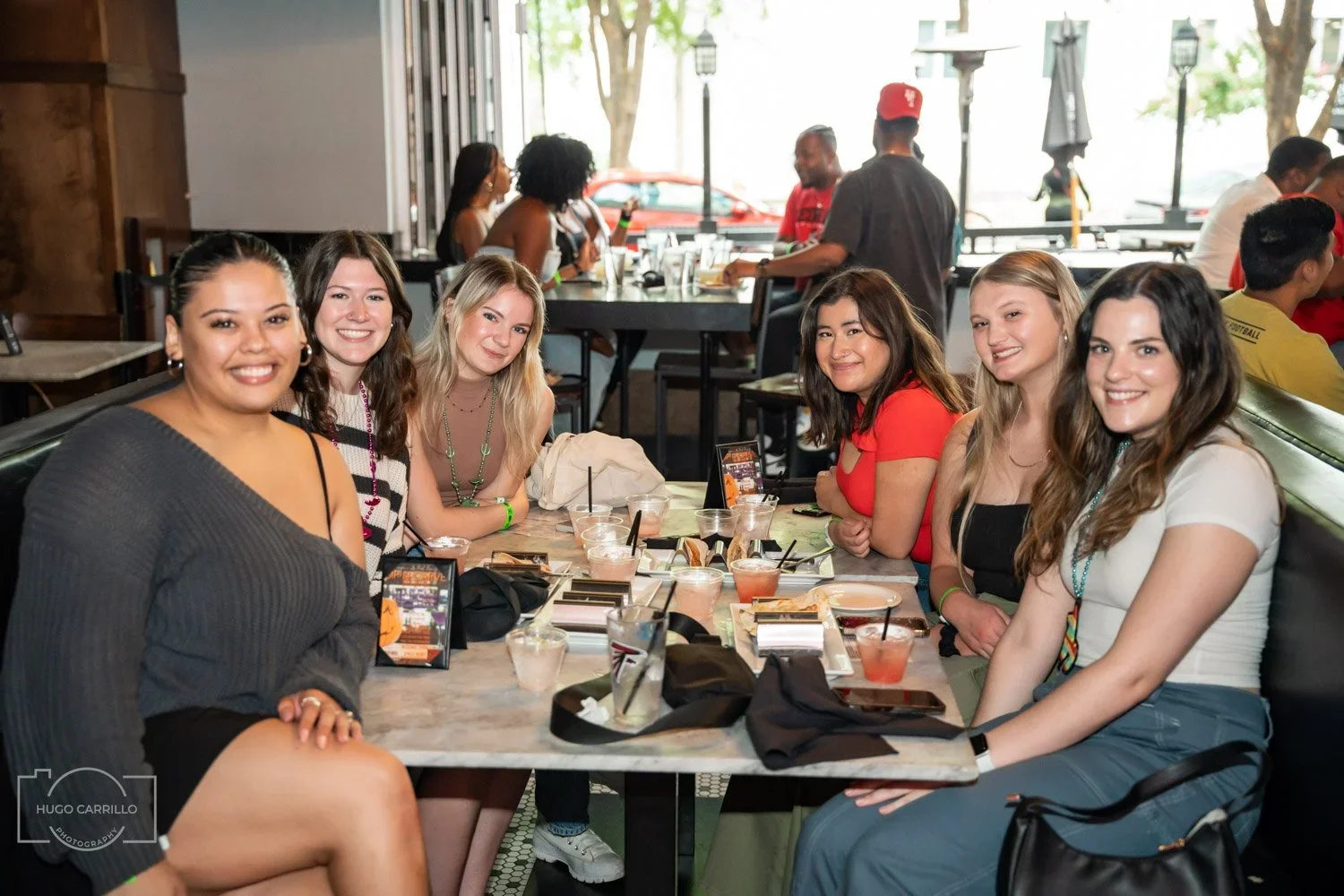 Group of seven young women sitting at a restaurant table, smiling at the camera, with drinks and food on the table.