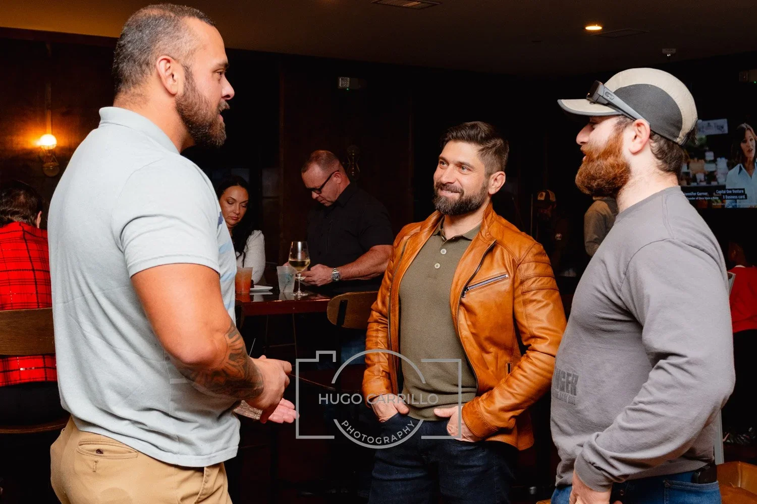 Three men conversing in a bar or restaurant, with others in the background, some holding drinks.