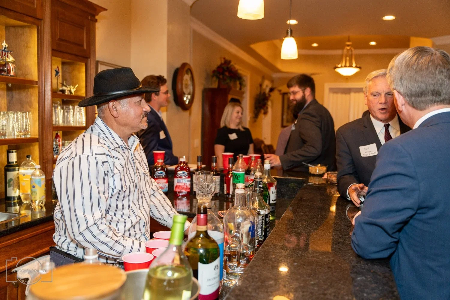 People socializing at a home bar, some dressed in suits, with bottles of alcohol, red cups, and glasses on the counter.