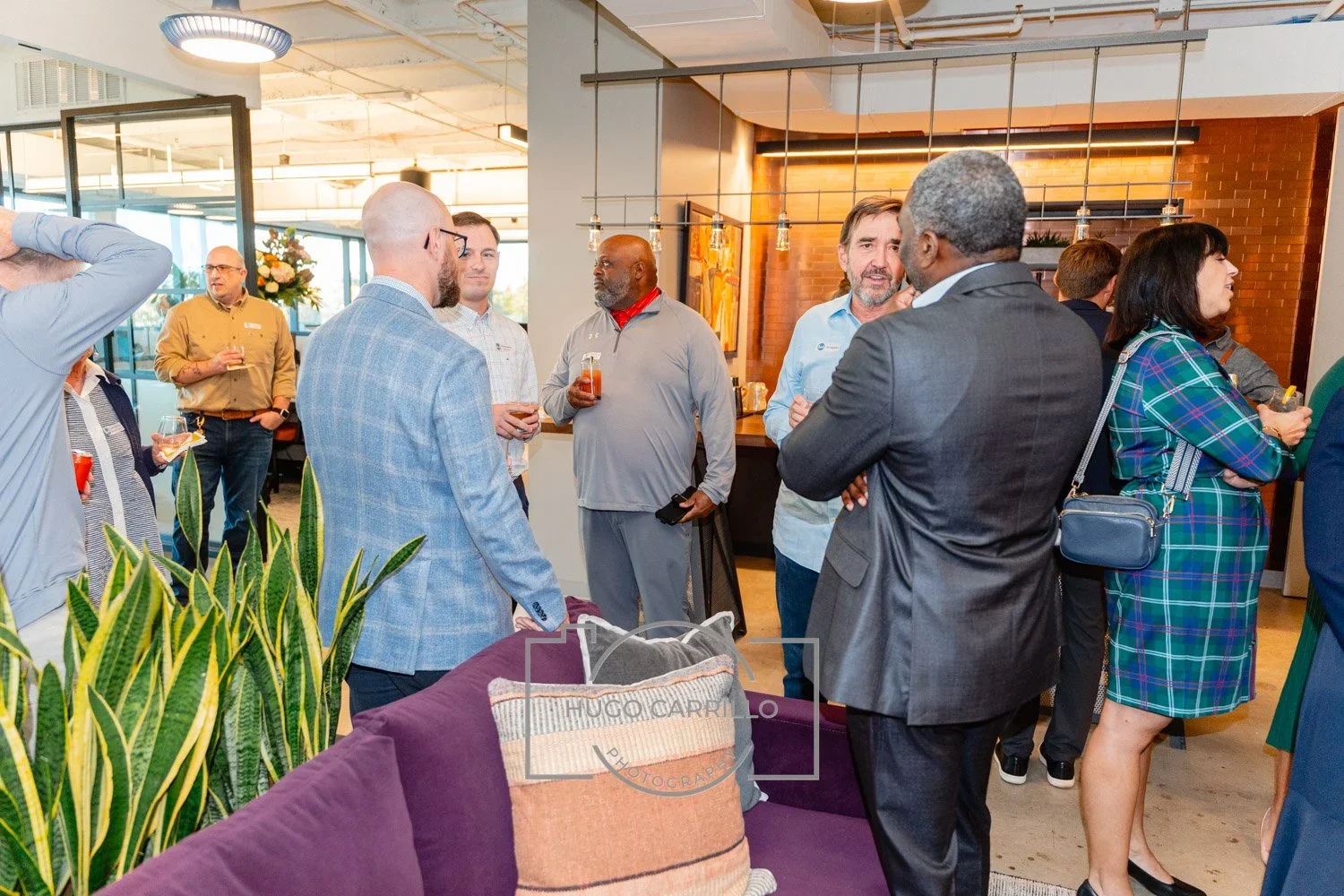 Group of diverse people socializing in a modern indoor setting with plants, artwork, and pendant lighting, some holding drinks and engaging in conversations.