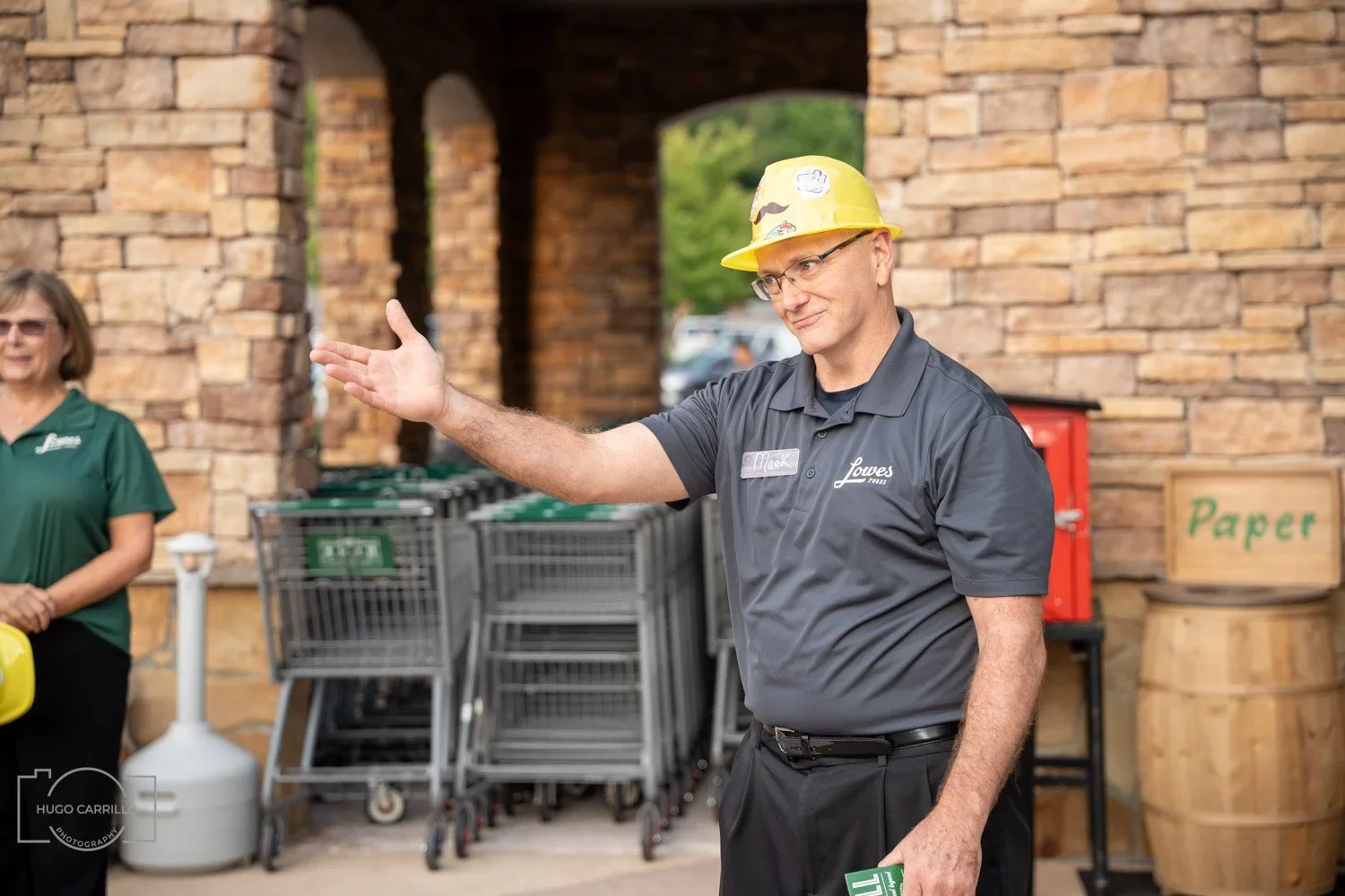 A man wearing a yellow hard hat, glasses, and a grey Lowe's shirt is gesturing with his right hand. A woman with sunglasses and a green shirt stands in the background near grocery carts. They are outside near a brick wall.