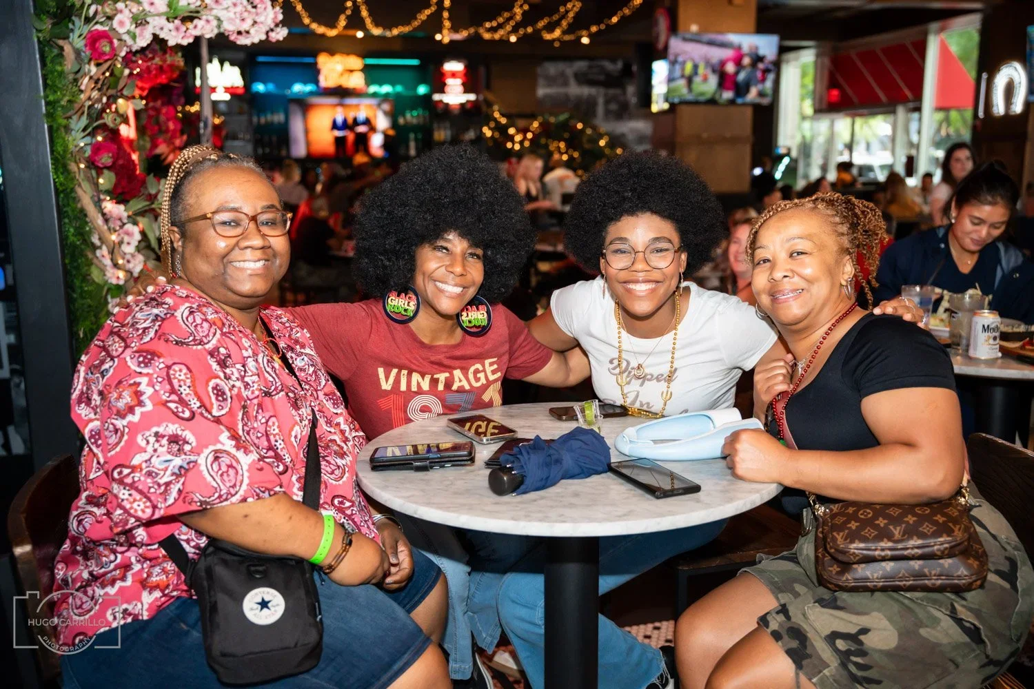 Four women sitting at a table in a lively restaurant or bar, smiling at the camera, with drinks and phones on the table, decorated with string lights and flowers.