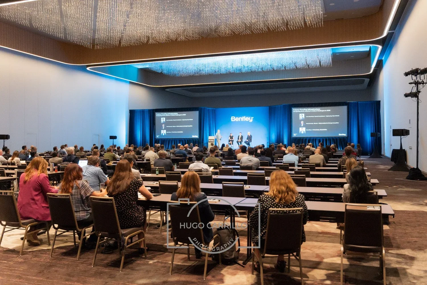 A large conference room with many attendees seated facing a stage with three speakers, illuminated by blue lighting and a large Bentley logo screen.