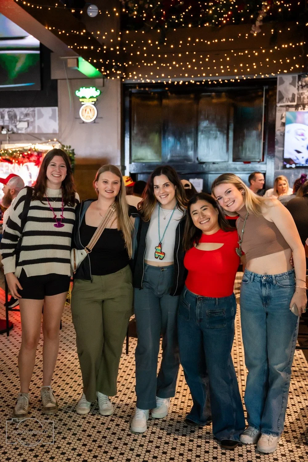 Five young women smiling and posing together in a lively, decorated bar or restaurant with string lights and a television in the background.
