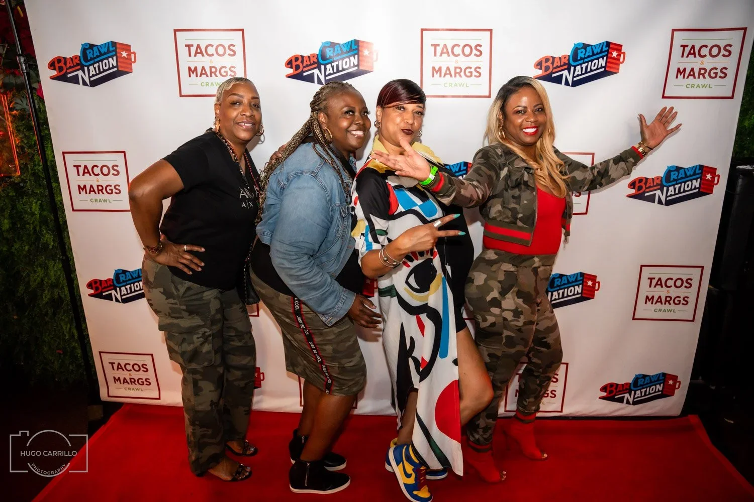 Four women posing on a red carpet in front of a backdrop with logos for Tacos & Margs Crawl and Bar Rawl Nation, smiling and making gestures.