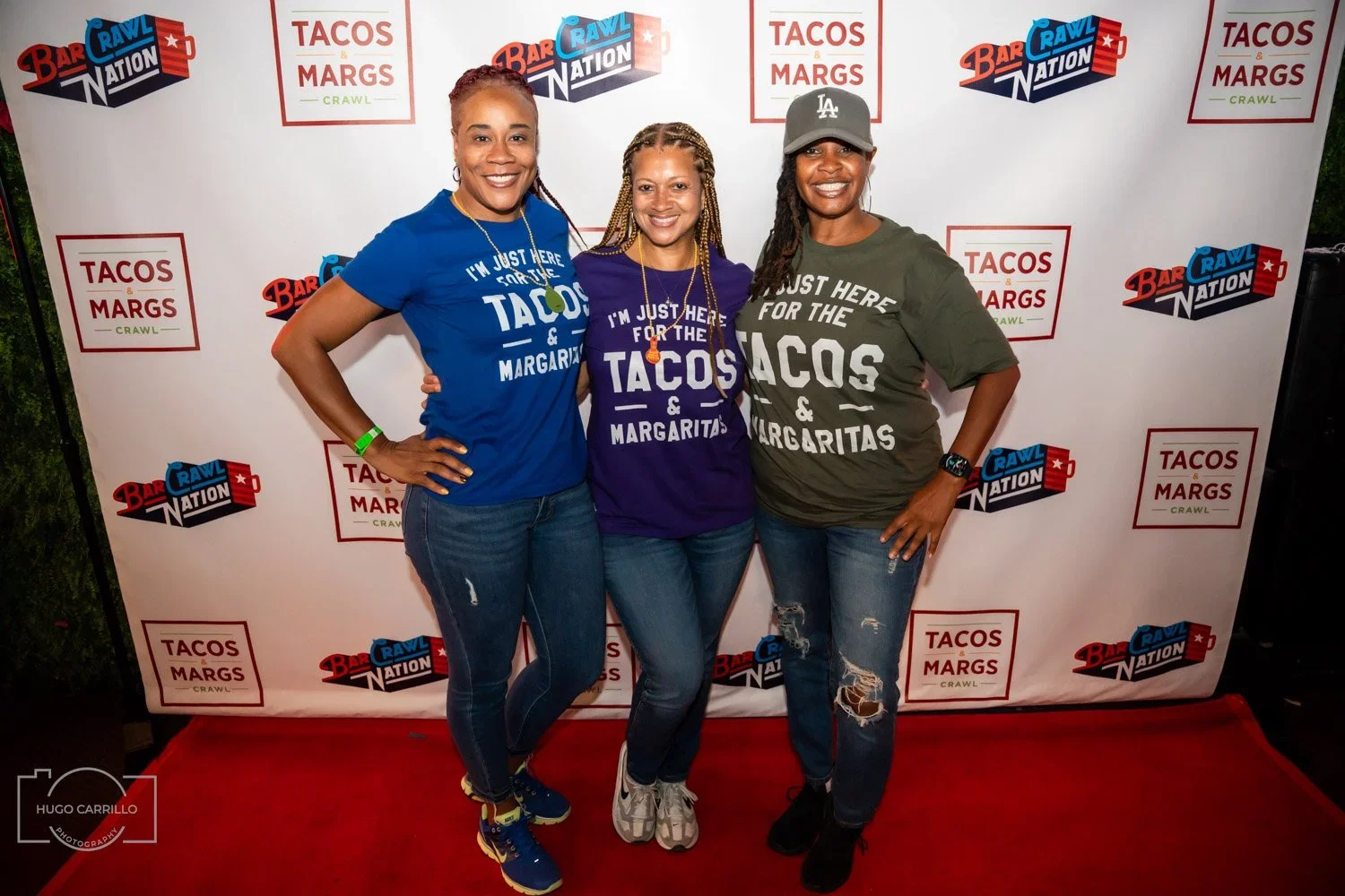 Three women standing in front of a backdrop with logos of Bar Rawl Nation and Tacos & Margaritas. They are smiling, wearing shirts that say 'I'm just here for the tacos & margaritas', with different colors and styles. The woman on the left is in a bl