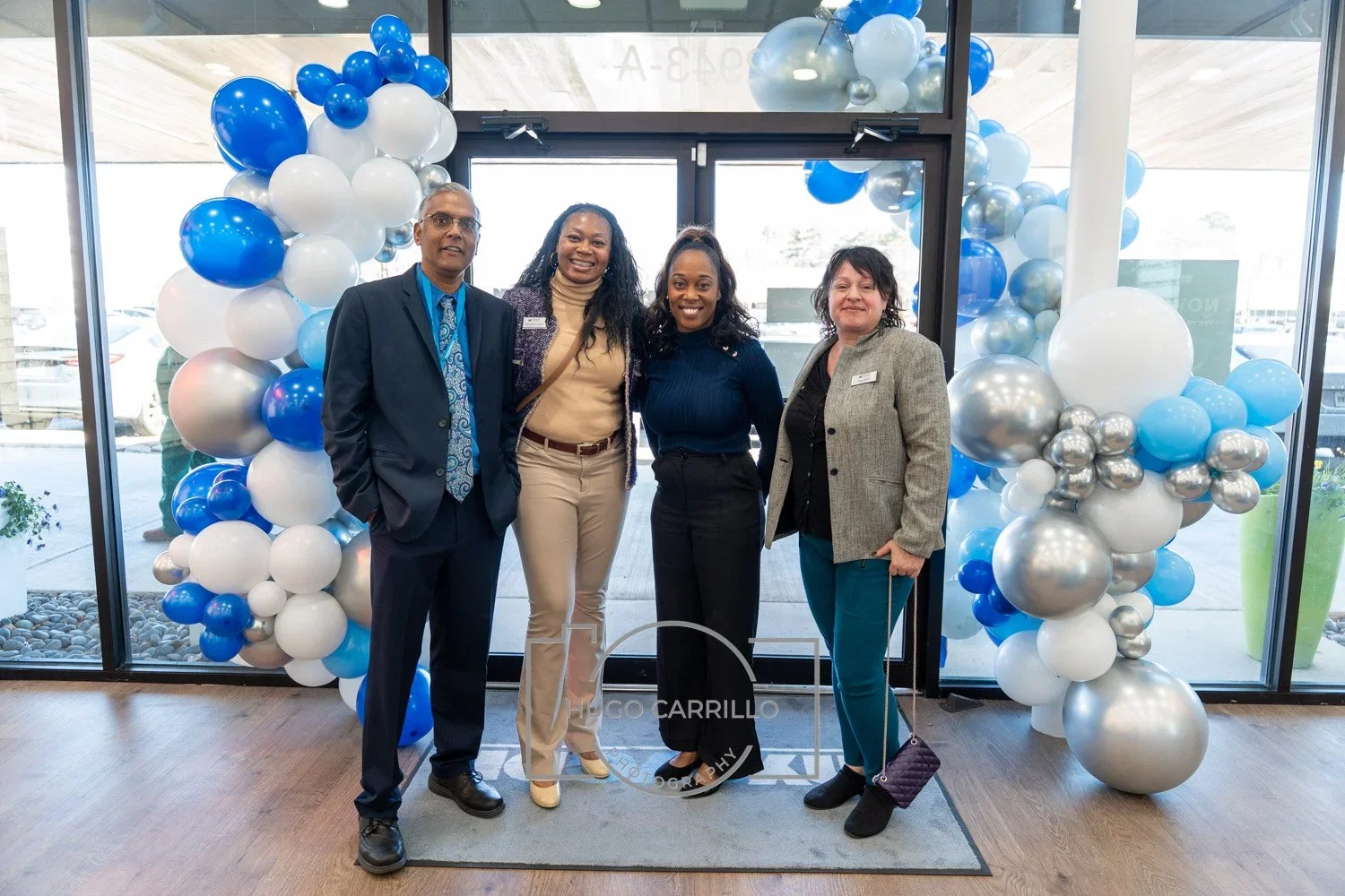 Four people standing together in front of a balloon arch, smiling, at an indoor event.