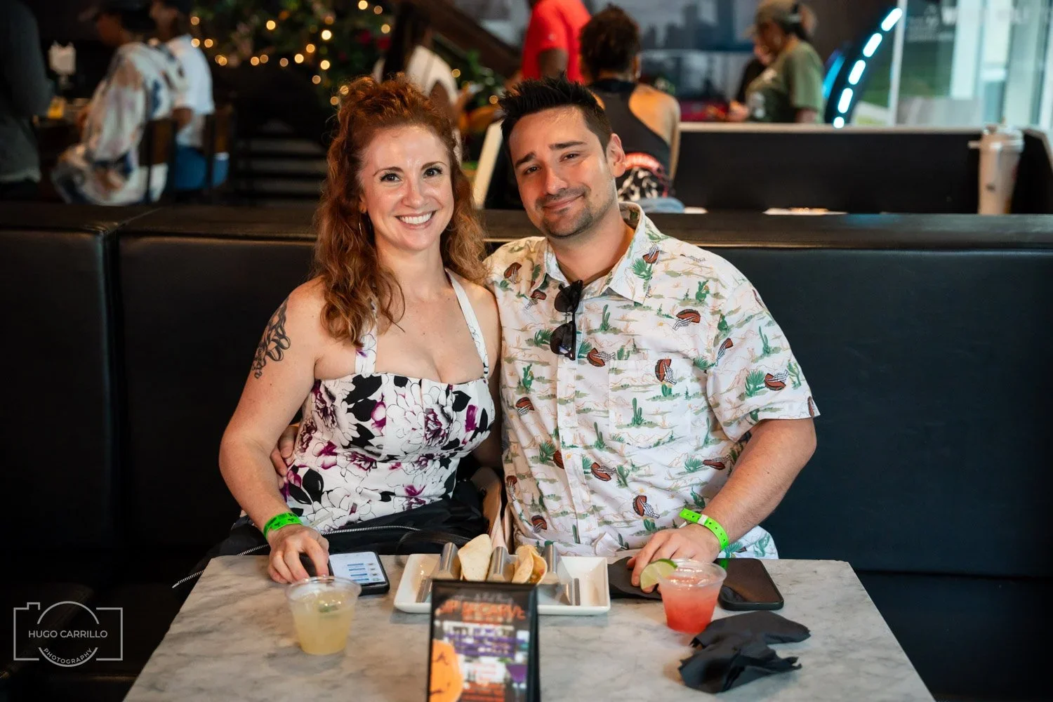 A smiling woman and man sitting at a table with drinks and food in a lively restaurant or bar, with people in the background and festive decorations.