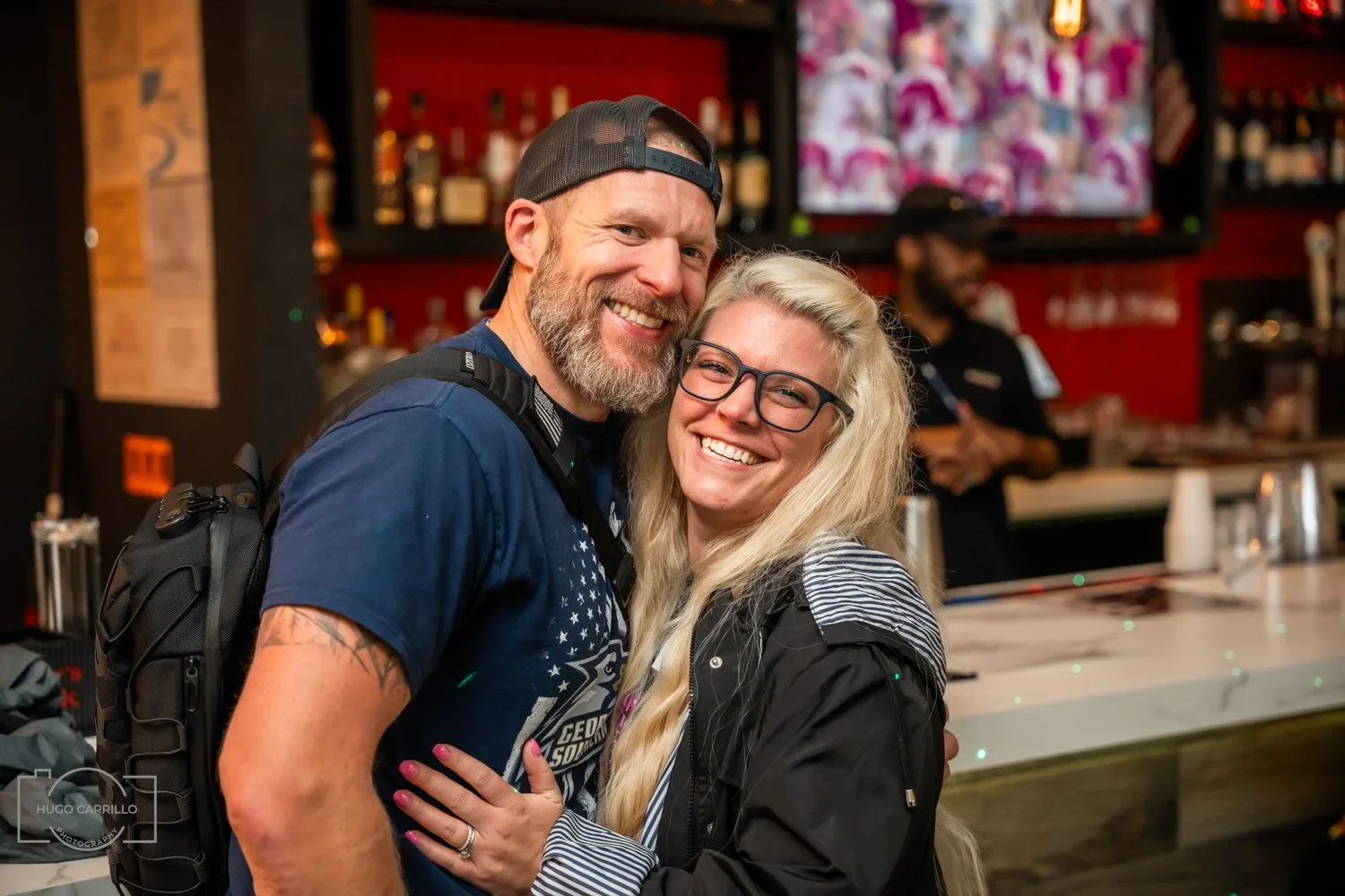A smiling man and woman embrace in a bar, with a bartender and bar equipment in the background.