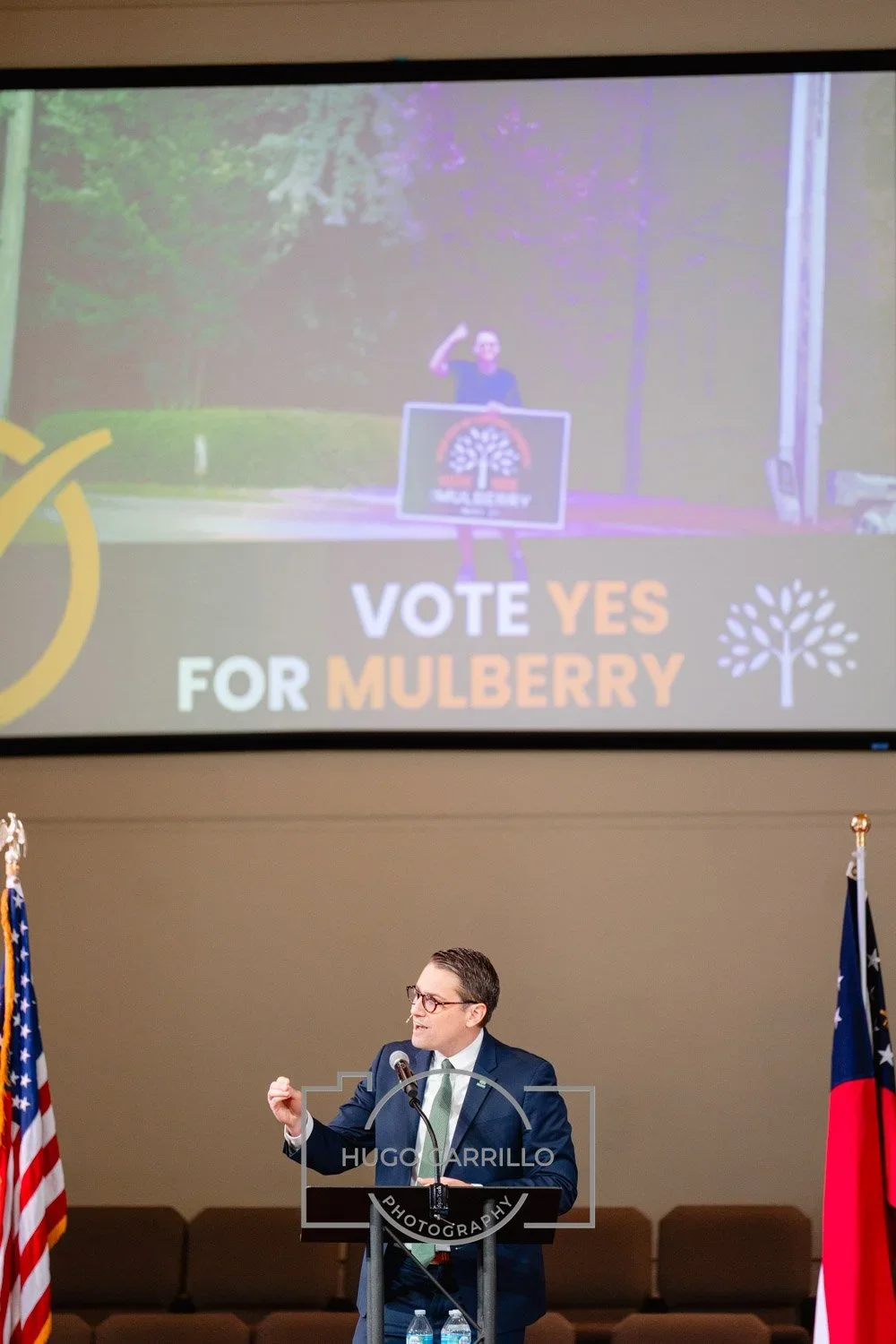 A man in a suit and glasses speaking at a podium in front of a large screen and two American flags. The screen shows a campaign advertisement with a hashtag and the text "VOTE YES FOR MULBERRY" along with an image of a person making a fist.
