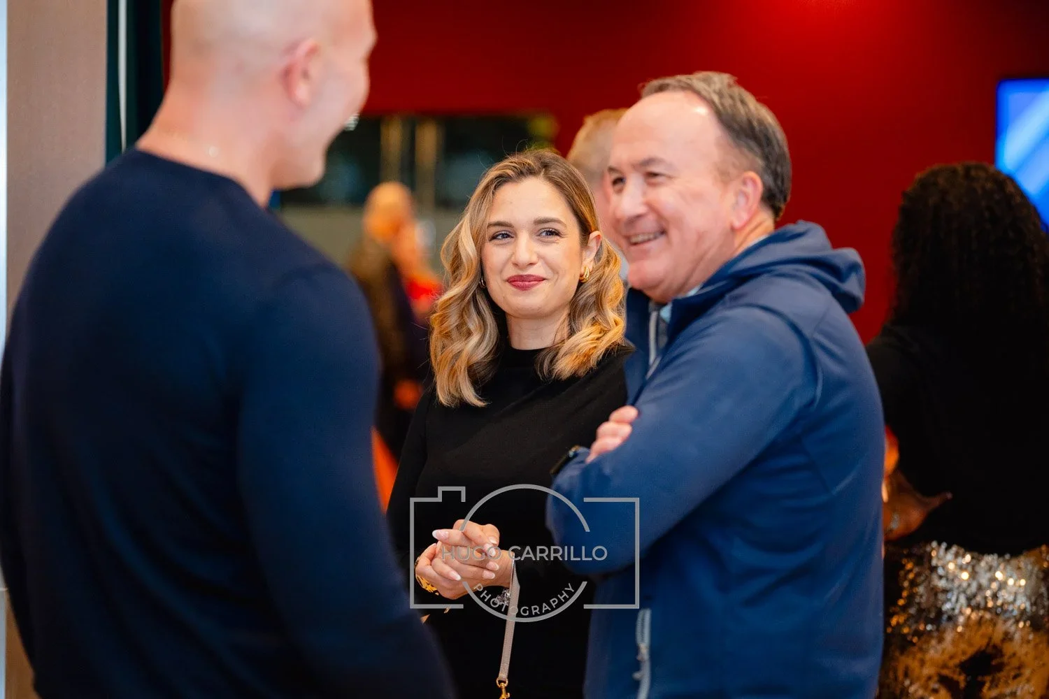 Three people talking at a social event, smiling and engaging in conversation, with a woman and a man in the foreground and another person in the background.
