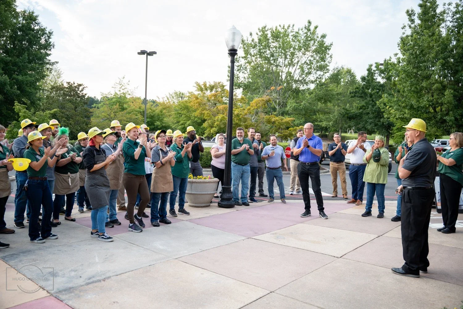 Group of people gathering outside, wearing yellow safety helmets, and applauding, with a person in blue speaking to them, in front of trees and parked cars.
