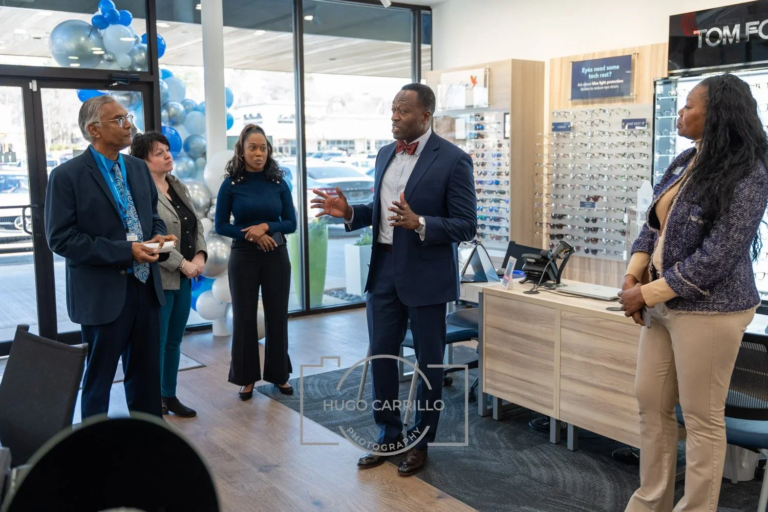 A man in a suit and bow tie is speaking to four women in a retail store, possibly an eyewear store, with display cases of glasses and large windows in the background.