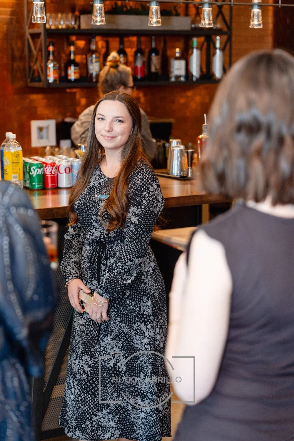 A woman smiling with long brown hair, wearing a black and white floral dress, standing in a bar or restaurant setting with drinks and snacks on the counter, and shelves of alcohol bottles behind her.