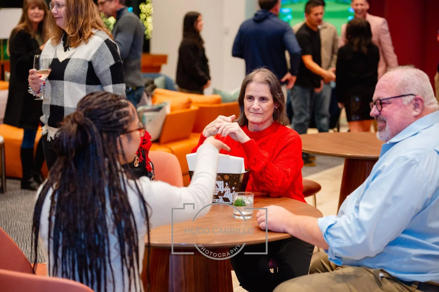 People sitting around a table chatting, with some standing and talking in the background at a social gathering.