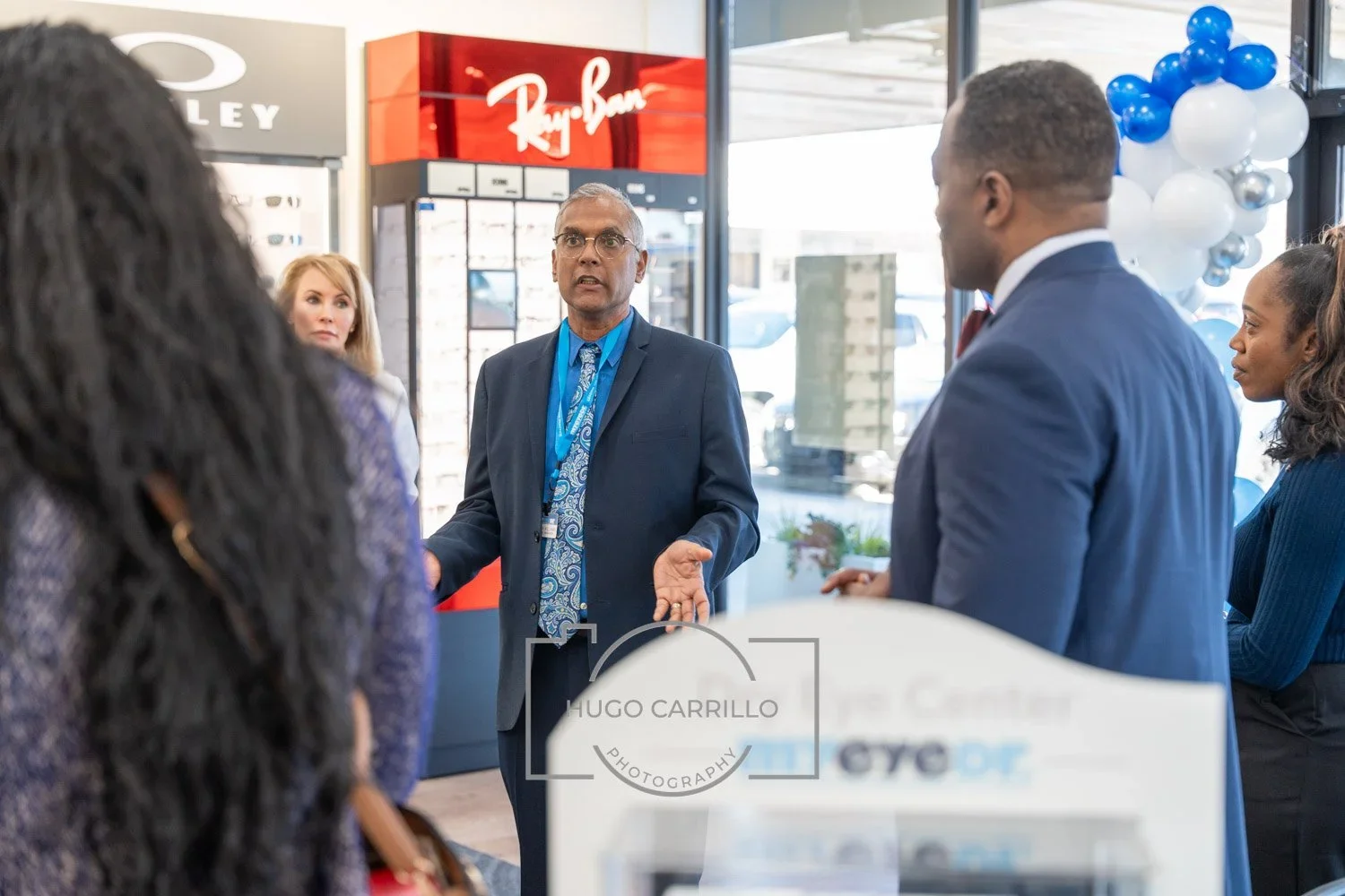 Man in a suit with glasses speaking to a group of diverse people in an optical store, with Ray-Ban signage and balloons in the background.