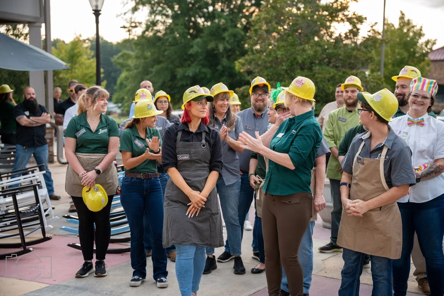 Group of people with yellow safety helmets, gathered outdoors, smiling, and talking to one another during daytime.