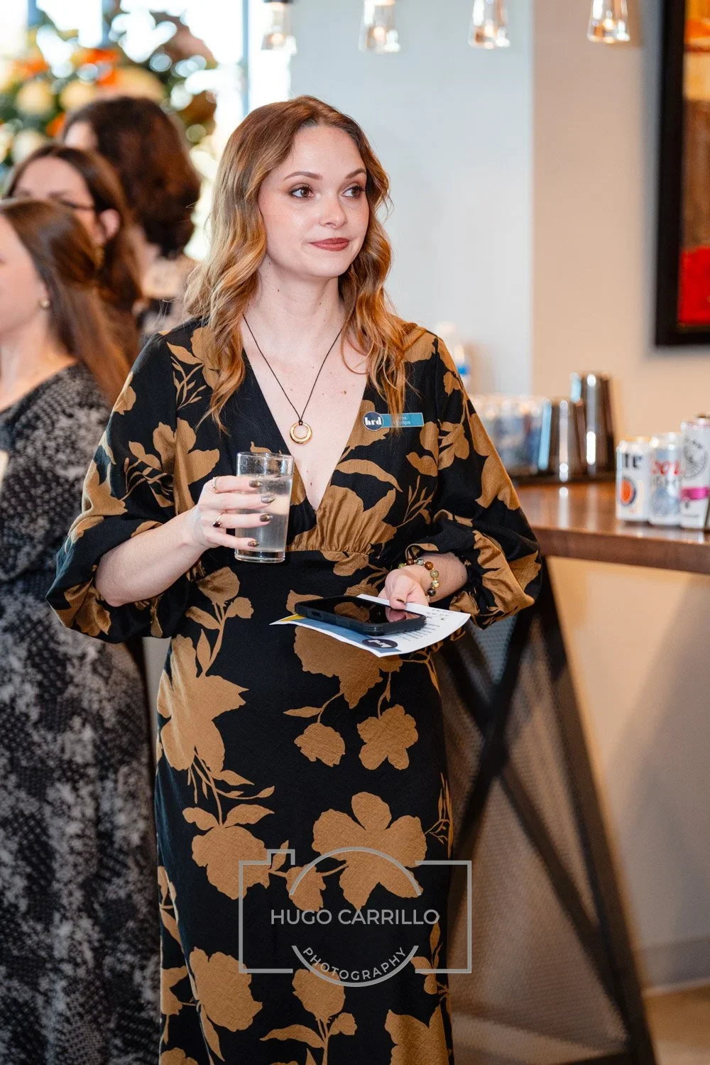 A woman with wavy, shoulder-length hair wearing a black dress with large tan floral patterns, holding a glass of water and some papers and a phone, at a social event or gathering.
