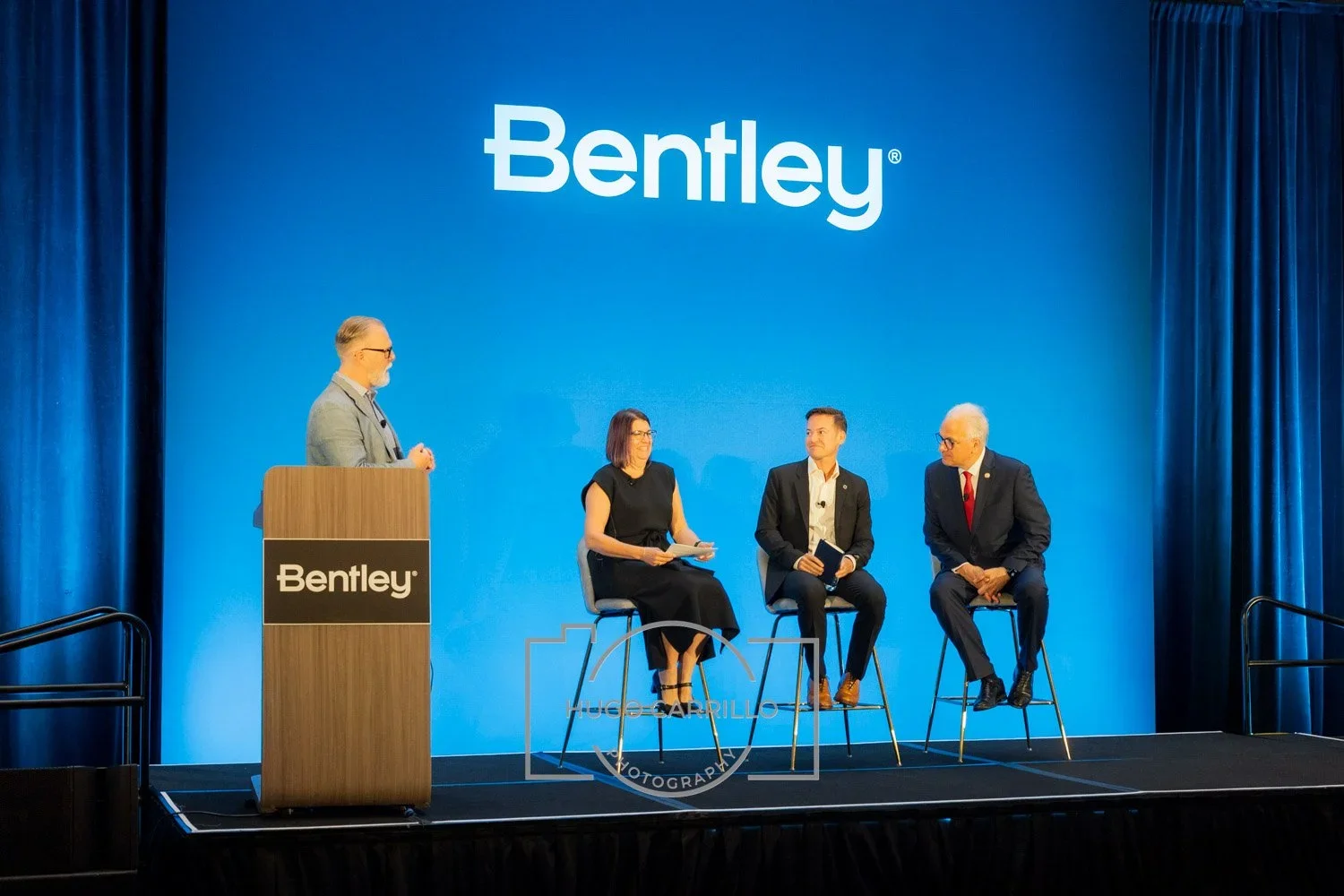 A panel discussion at a Bentley event with four people on stage, three sitting on stools and one standing at a podium, in front of a large blue background with the Bentley logo.