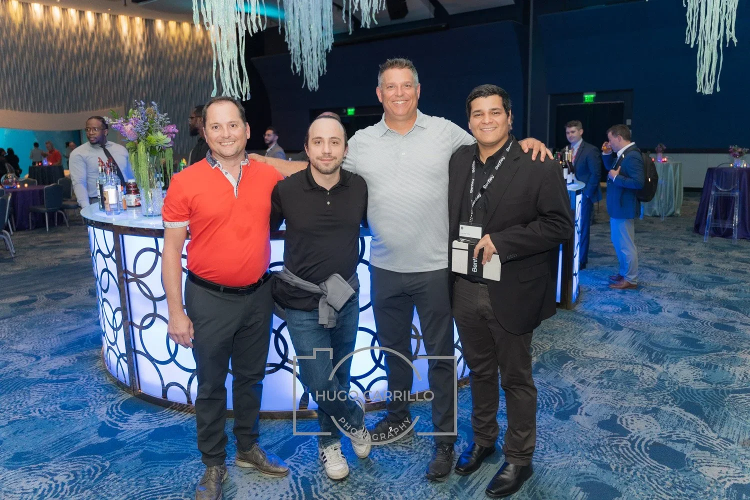 Four men standing together smiling at a social event with a decorated bar counter in the background.