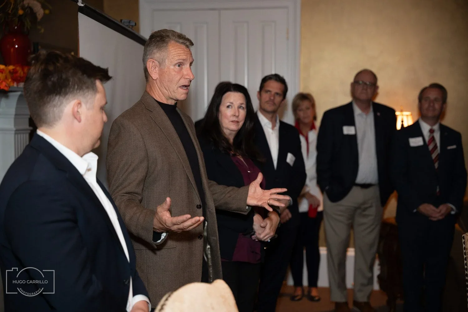 Man in brown blazer speaking to a group of people in business attire during a meeting or event