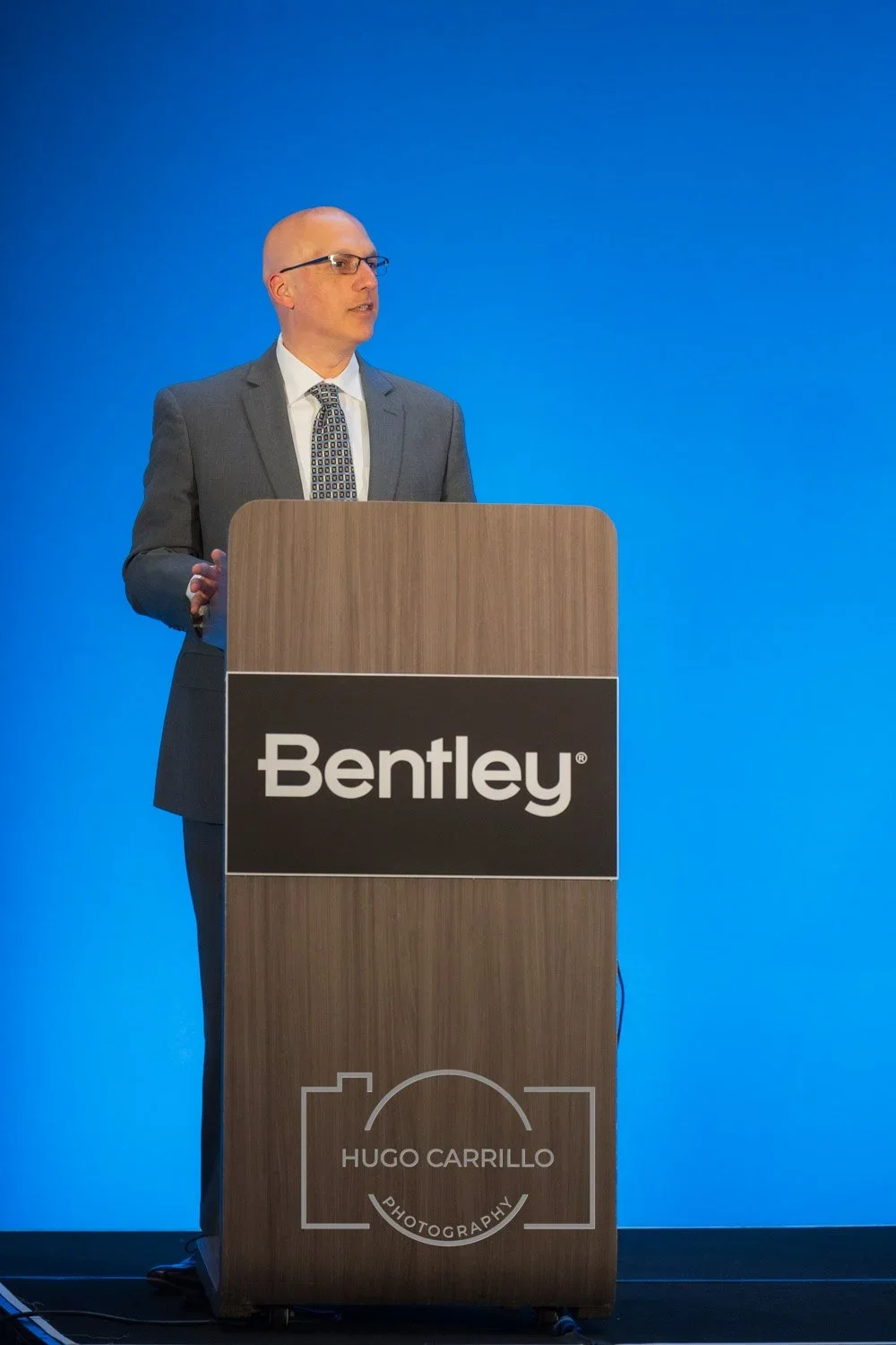 A man in a gray suit and glasses standing at a wooden podium with a Bentley logo, speaking at an event with a blue background.