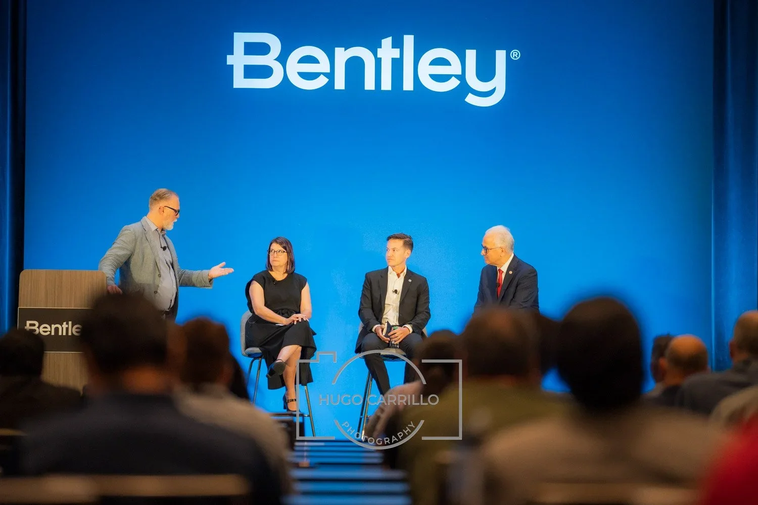 A panel discussion at a Bentley event with four people on stage, sitting on chairs facing the audience, with a large blue backdrop and the Bentley logo above them.