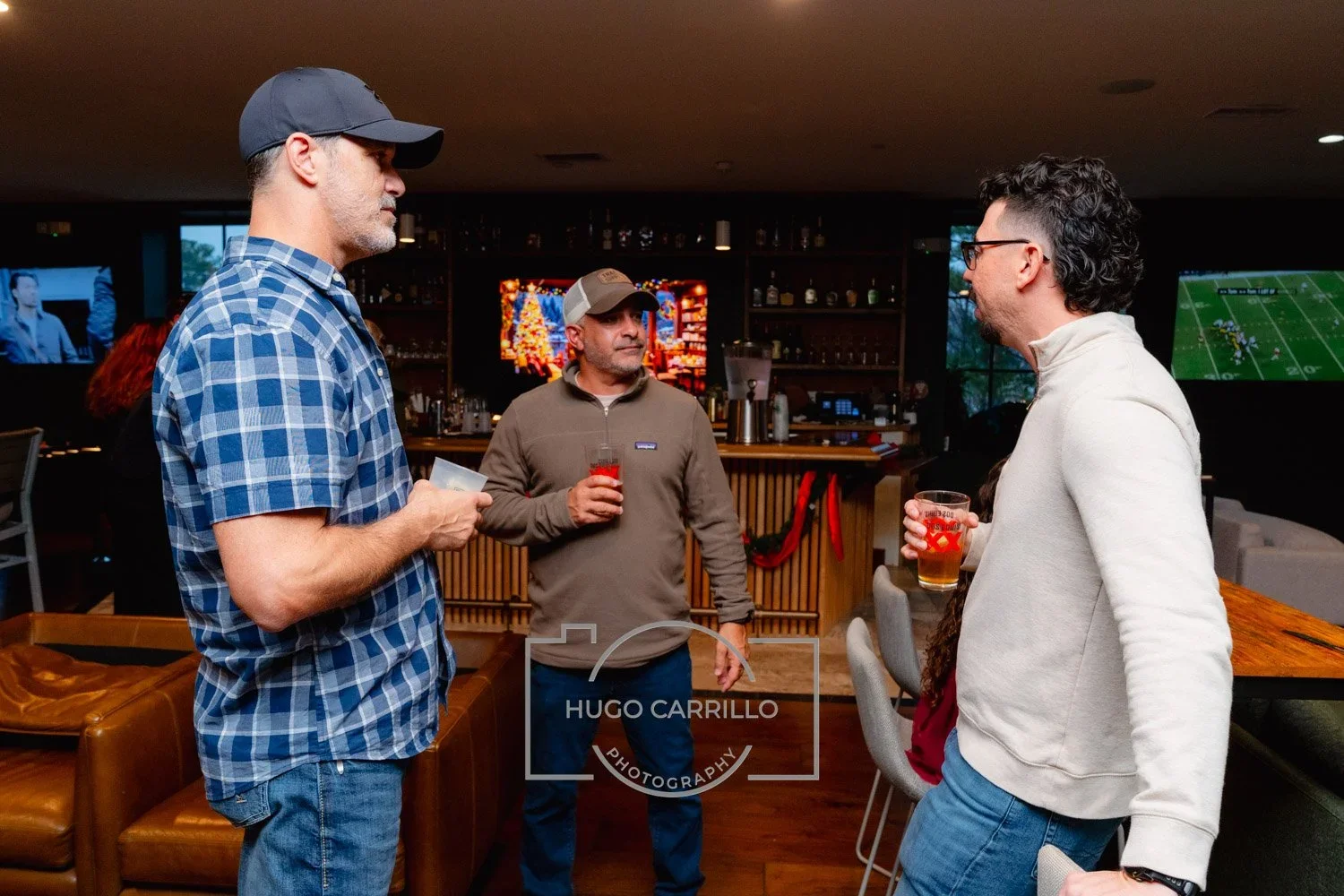 Three men are conversing at a bar, each holding a drink. The man on the left wears a blue checkered shirt and a dark hat, the man in the middle wears a brown zip-up jacket and a baseball cap, and the man on the right wears a white quarter-zip sweater