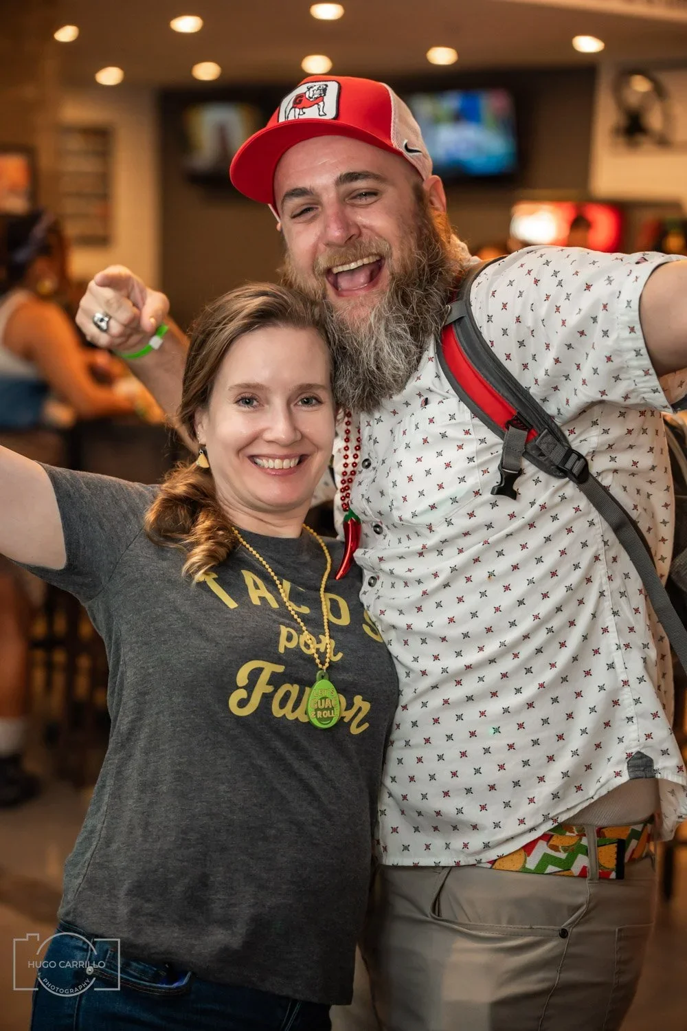 A man and woman smiling and celebrating in a busy indoor setting, with the woman wearing a 'Tacos for Farmer' t-shirt and the man wearing a patterned shirt, backpack, and a red and white cap.