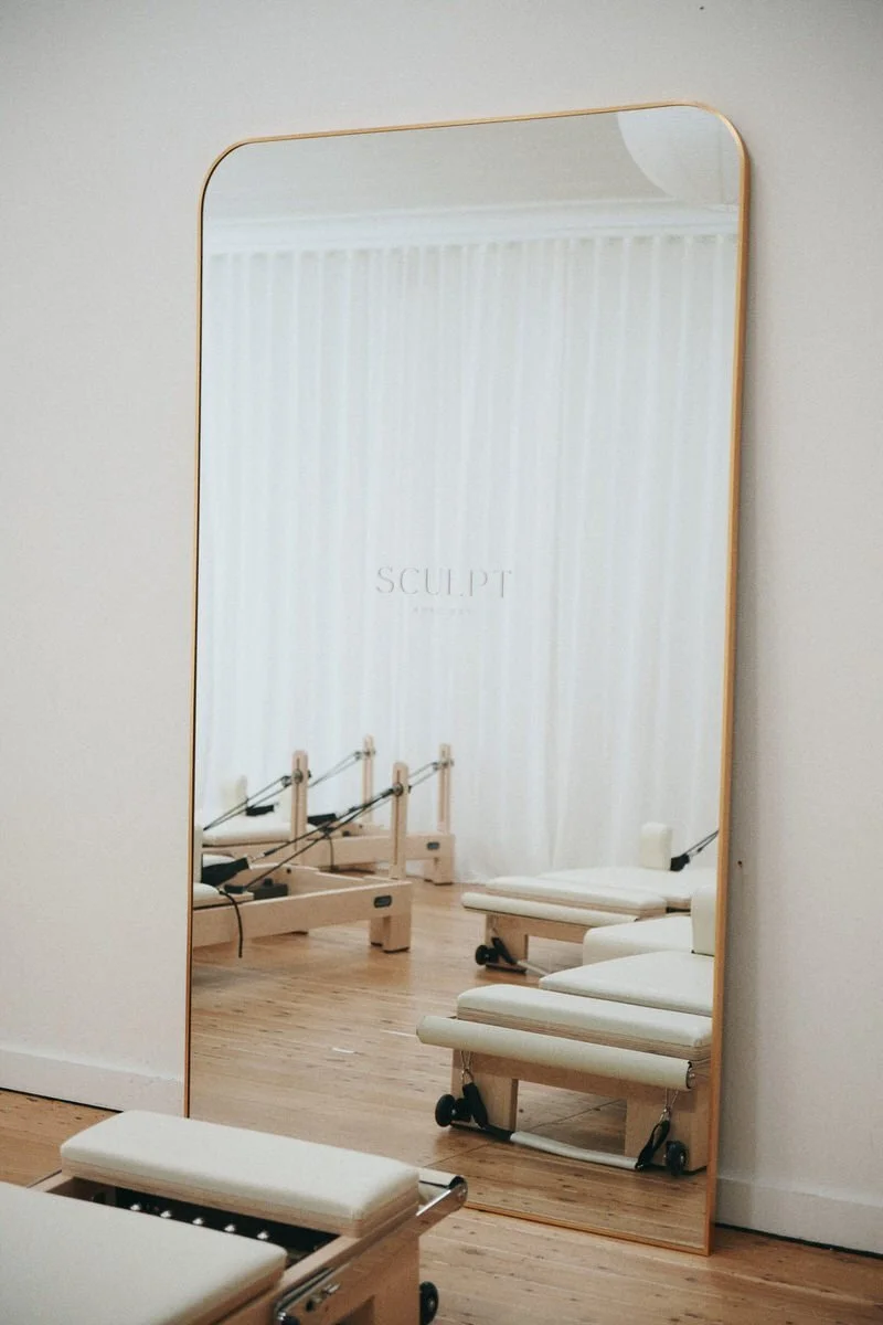 A gold-framed mirror with SCULPT Rose Bay etched on it, reflecting the reformers