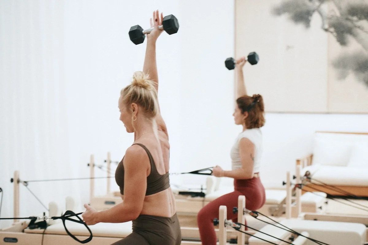 Two women raising dumbbells overhead on the reformers