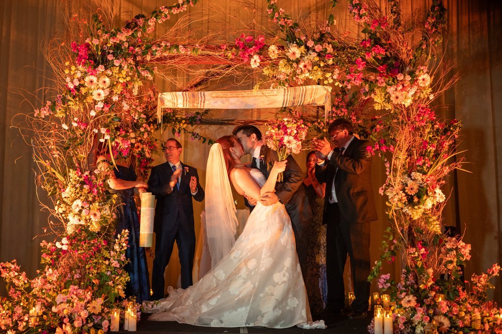 Sharon y Josh se besan bajo la jupá de flores rosadas y naranjas